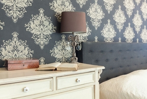 A bedroom with grey patterned wallpaper, a headboard with grey fabric, a cream dresser, and a lamp.