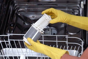Someone wearing yellow rubber gloves removing and cleaning a dishwasher filter.