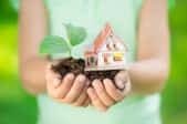 A child holding a pile of soil and a small house next to a leafy plant. 