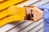 gloved hand painting yellow paint on a white exterior wall with a small brush