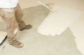 A professional painter uses a roller to spread white paint over a concrete floor.