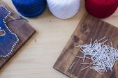 A trio of string in red, white, and blue colors next to string art and a pile of nails on a wood board. 