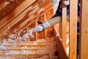 A worker spraying blown fiberglass insulation between attic trusses.