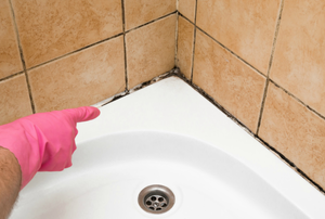 person with a pink glove pointing toward the mildew along the caulk around the bathtub