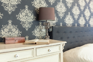 A bedroom with grey patterned wallpaper, a headboard with grey fabric, a cream dresser, and a lamp.