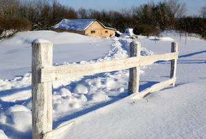 A snow fence surrounded by snow with a house in the background.