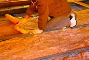 A man installing glass-wool insulation in between joists in an unfinished room.