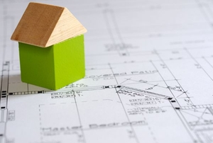 A green and wood toy block house sitting on some blueprints.