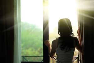 A woman opening up curtains in the morning with sunshine streaming through. 