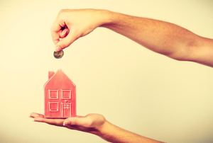 Someone holding a small red house piggy bank with a coin over it.