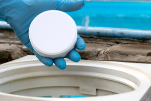 gloved hand dropping large chlorine tablet into skimmer to treat pool water