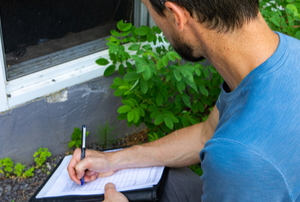 man inspecting exterior of basement window with clipboard and pen
