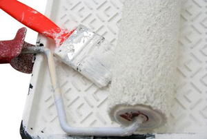 A paint roller and brush laying in a tray covered with white paint.