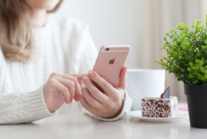 A woman using an iphone on a white tabletop with a green plant and a slice of cake. 