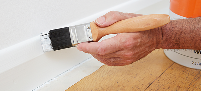 A man painting a baseboard with white paint. 