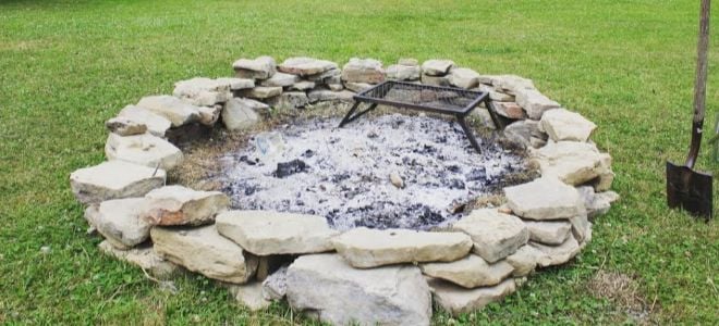 A fire pit in a grassy lawn with a wooden barn and trees in the background. 