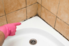 person with a pink glove pointing toward the mildew along the caulk around the bathtub
