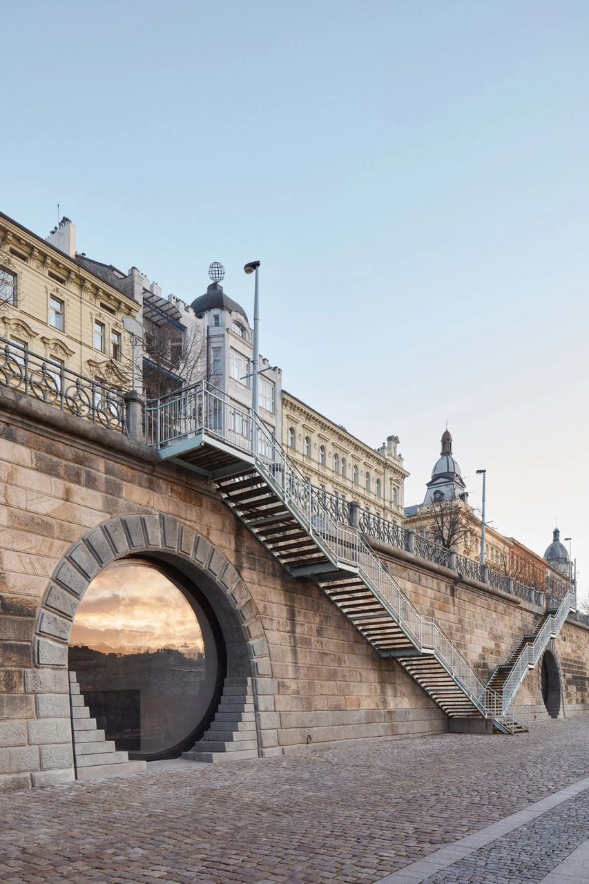 View of a windowed Vltava River vault underneath a metal stairway leading down to the embankment. 