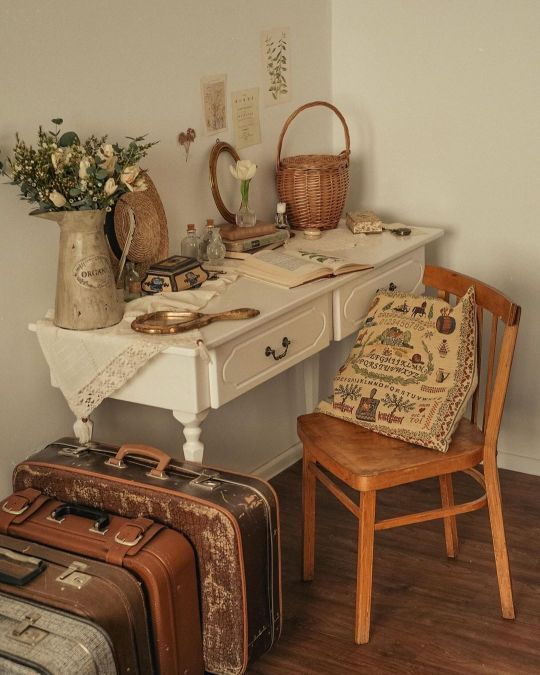 A beautiful example of cottagecore home decor, complete with a rustic white desk, vintage suitcases, and dried flowers arranged tastefully in old pots.
