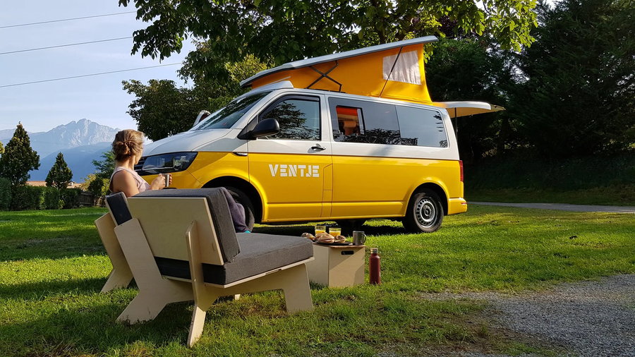 Woman relaxes on fold-out chairs outside her Ventje VW campervan, with the pop top fully up. 