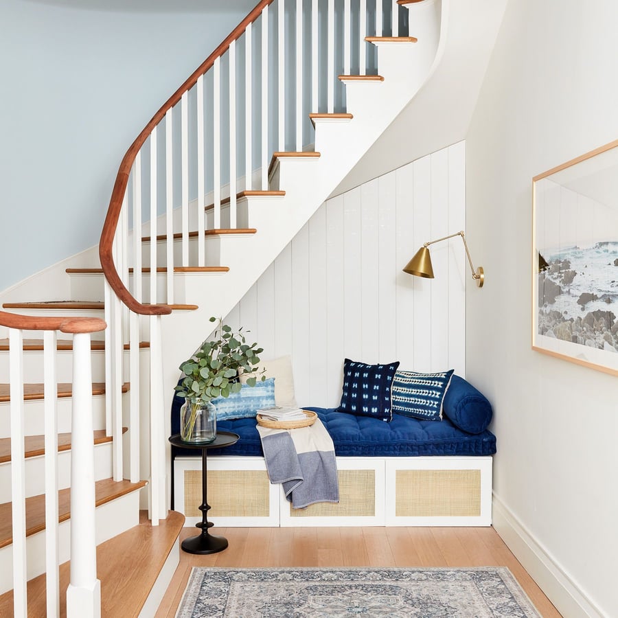 Stylish throw pillows and a gold wall fixture adorn the cozy area under the 2020 Real Simple Home's main staircase.