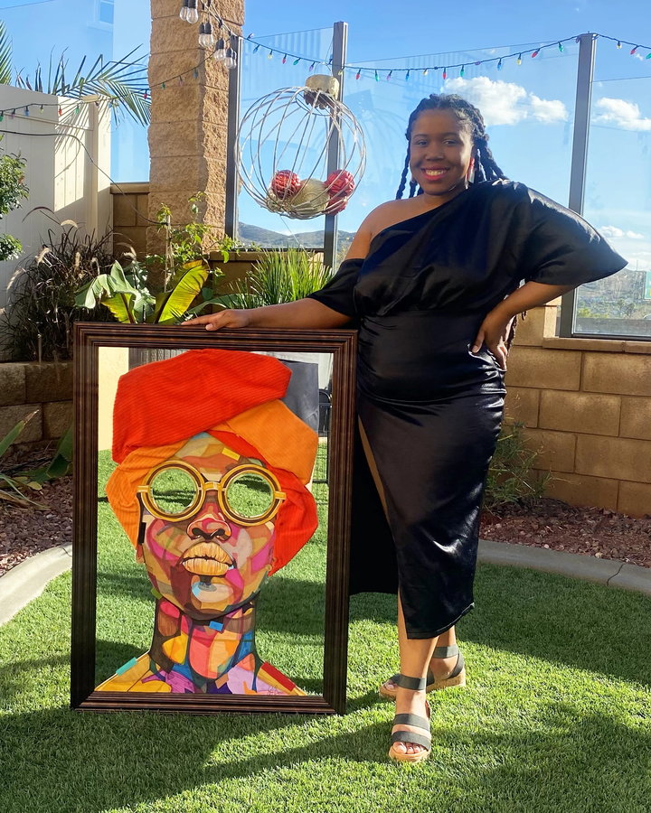 Domonique Brown holds up a mirrored DomoINK Art piece depicting a Black woman wearing a headwrap and sunglasses.