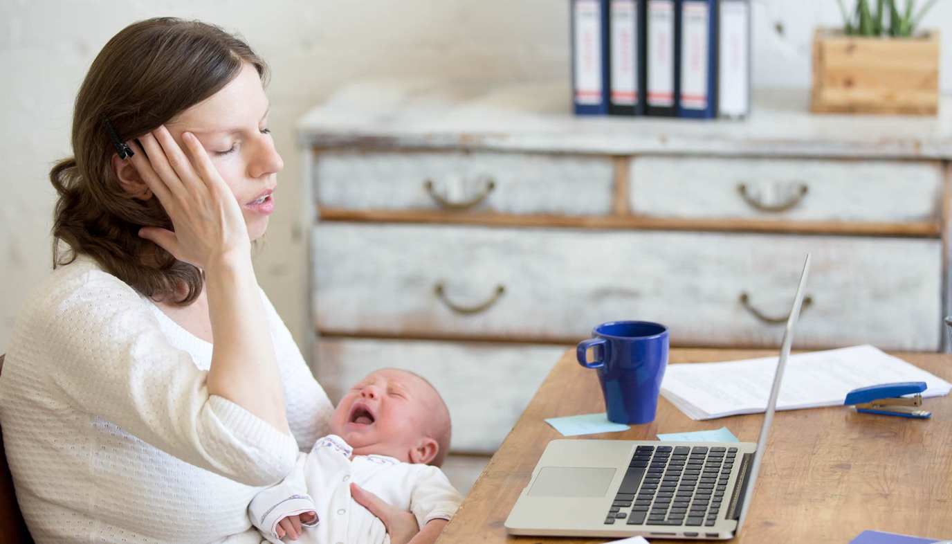 new mom touching her head in pain while using a laptop and holding a baby