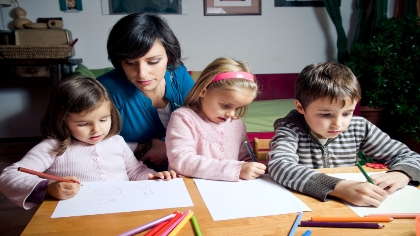 A woman watches over three children who are coloring pictures on paper. 